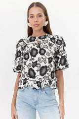 Model wears white, short-sleeve blouse with black, floral lace and buttoned neckline.