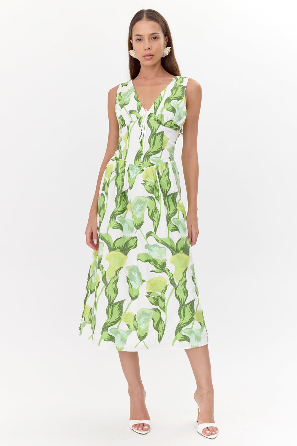 Model wears a sleeveless midi dress. The dress has a white base with a green floral print. The dress features a V-neckline, a corset-shaped bodice, and a tie front detail.