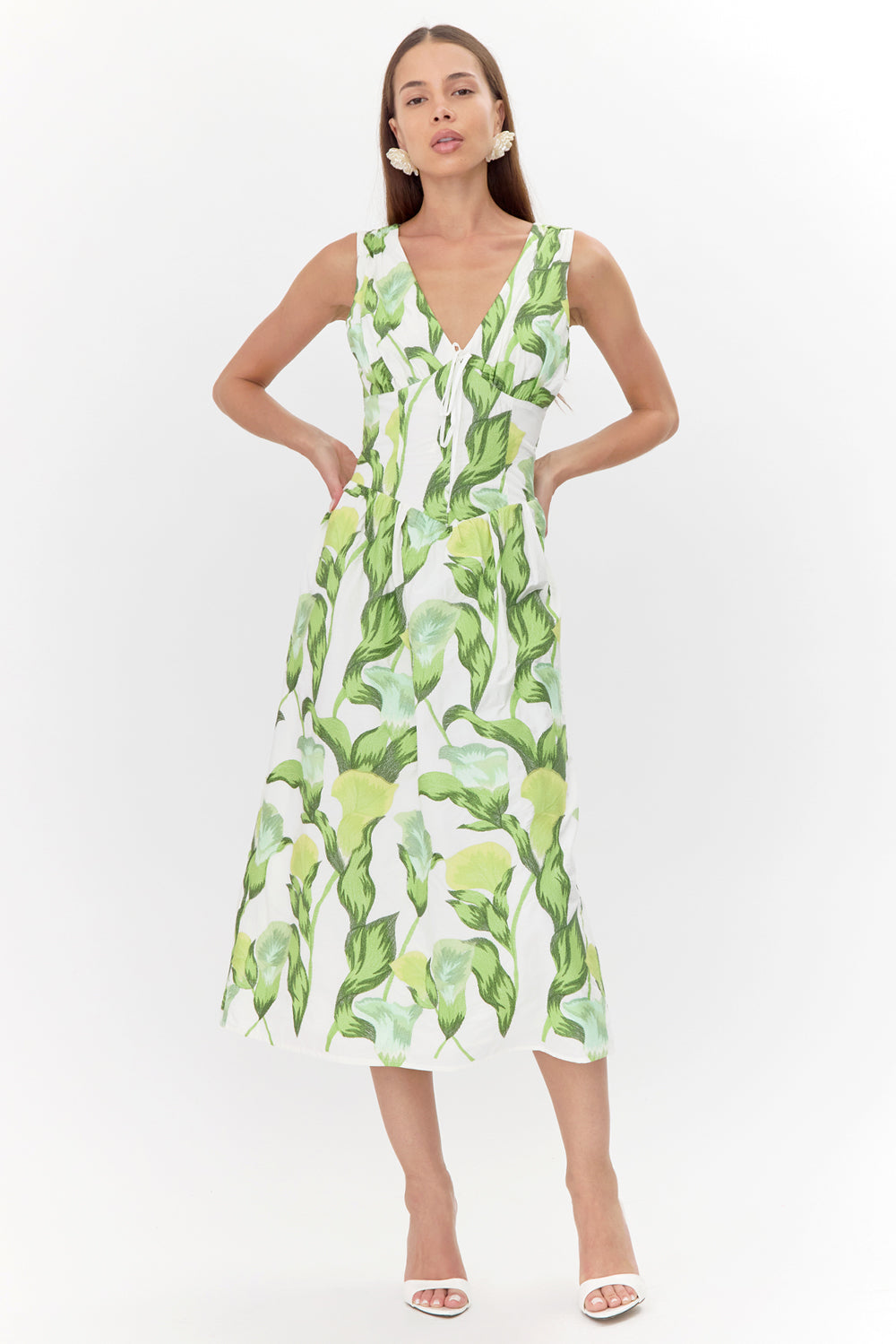 Model wears a sleeveless midi dress. The dress has a white base with a green floral print. The dress features a V-neckline, a corset-shaped bodice, and a tie front detail.