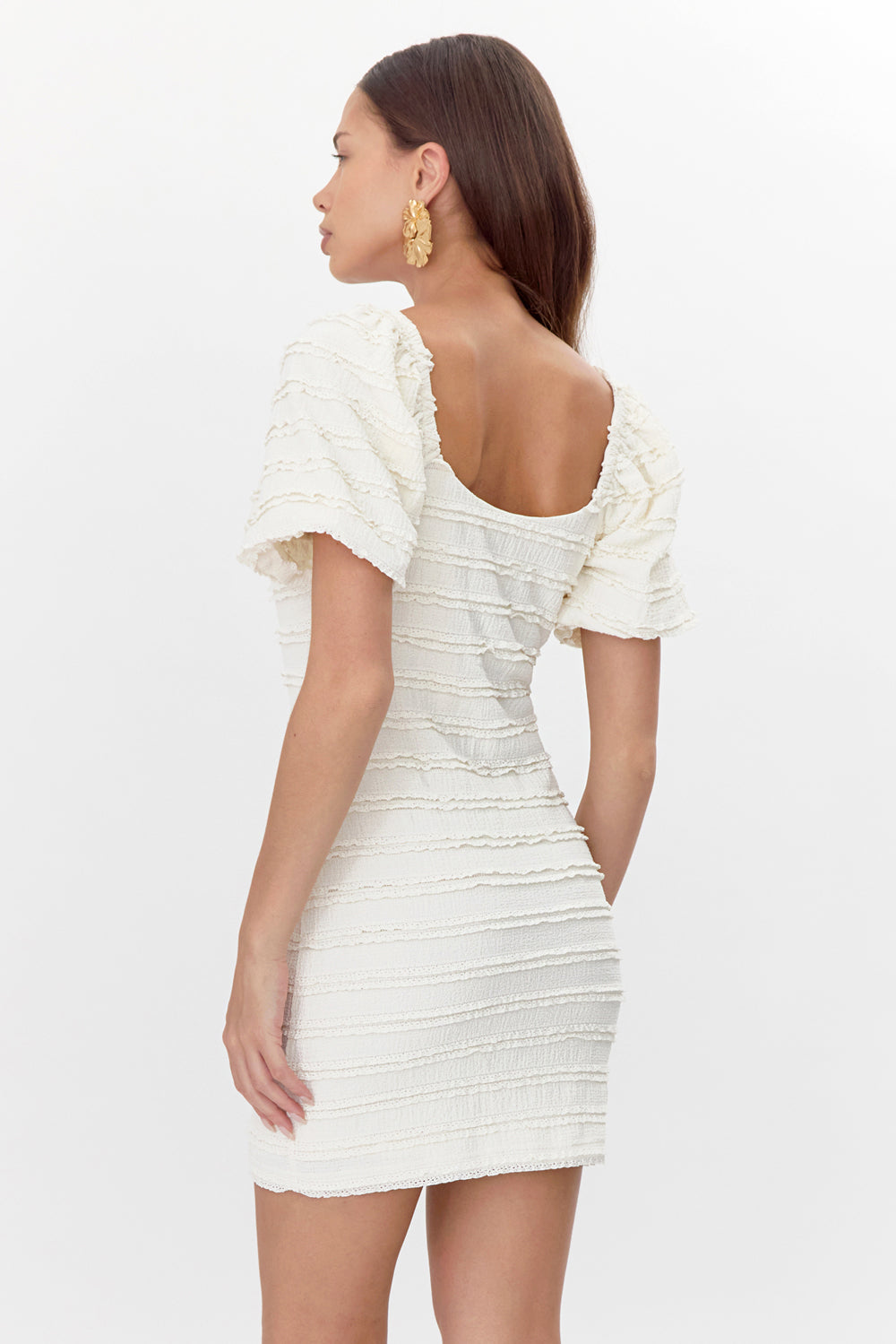 Back view of model wearing white, form-fitting mini dress featuring short sleeves and a small, ruffled texture. The back has a deep drop, exposing the top half of the model's back.