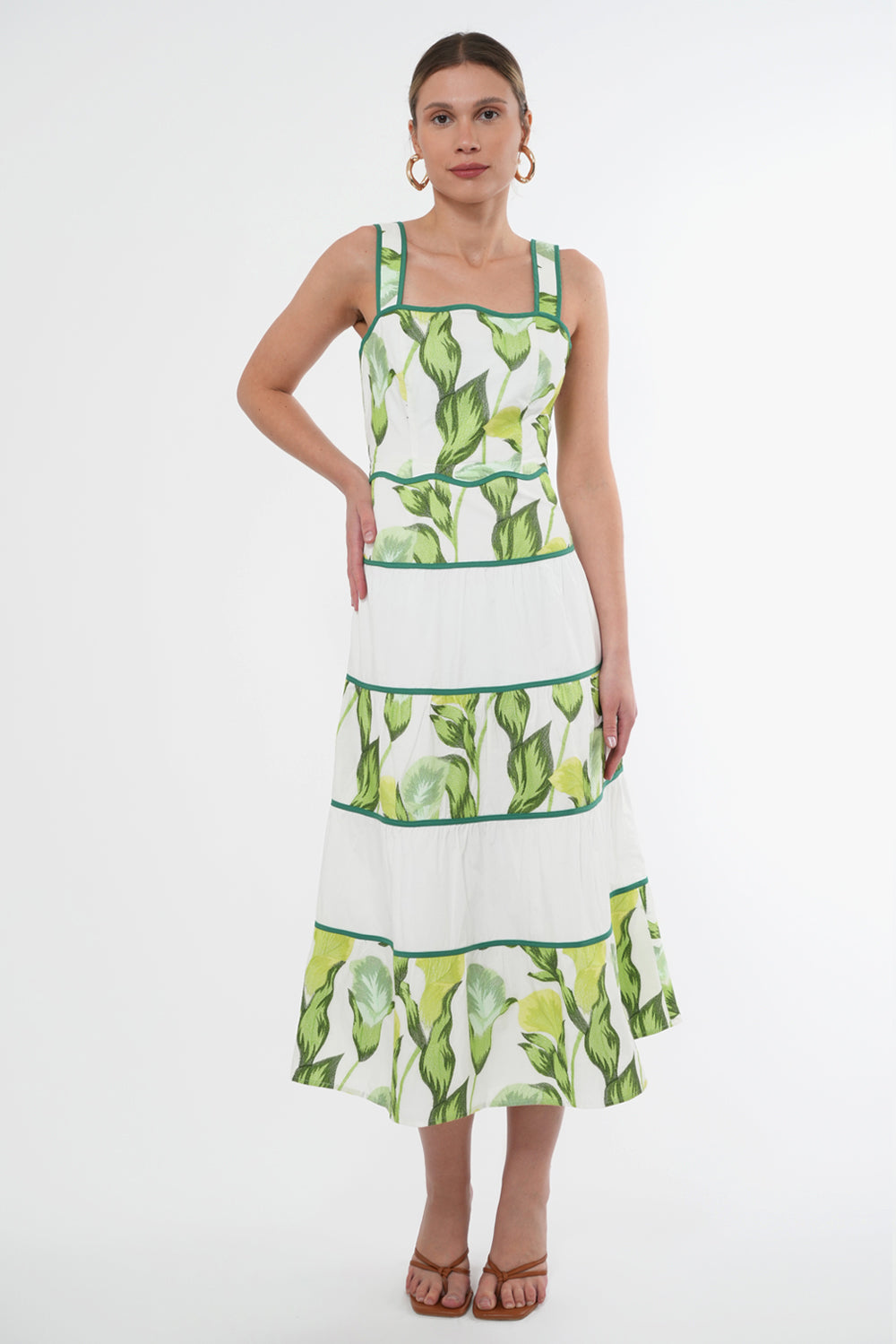 Model wears a sleeveless midi dress the dress features a green floral print on top of a white base. The neckline and waistline is scalloped. The skirt has contrasting, white tiers.