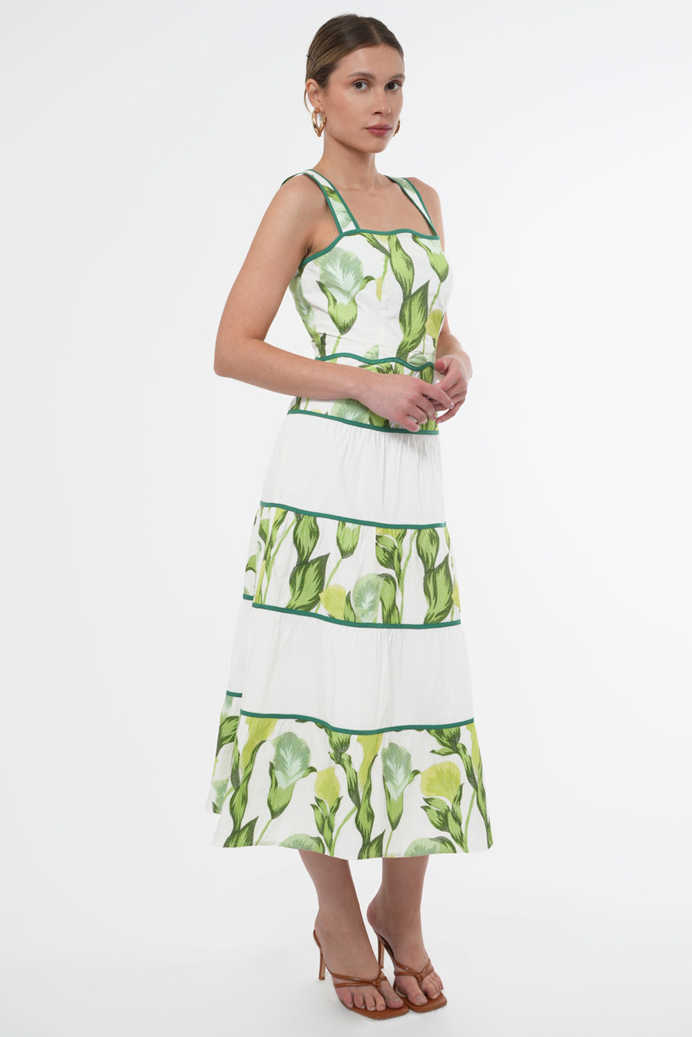 Model wears a sleeveless midi dress the dress features a green floral print on top of a white base. The neckline and waistline is scalloped. The skirt has contrasting, white tiers.