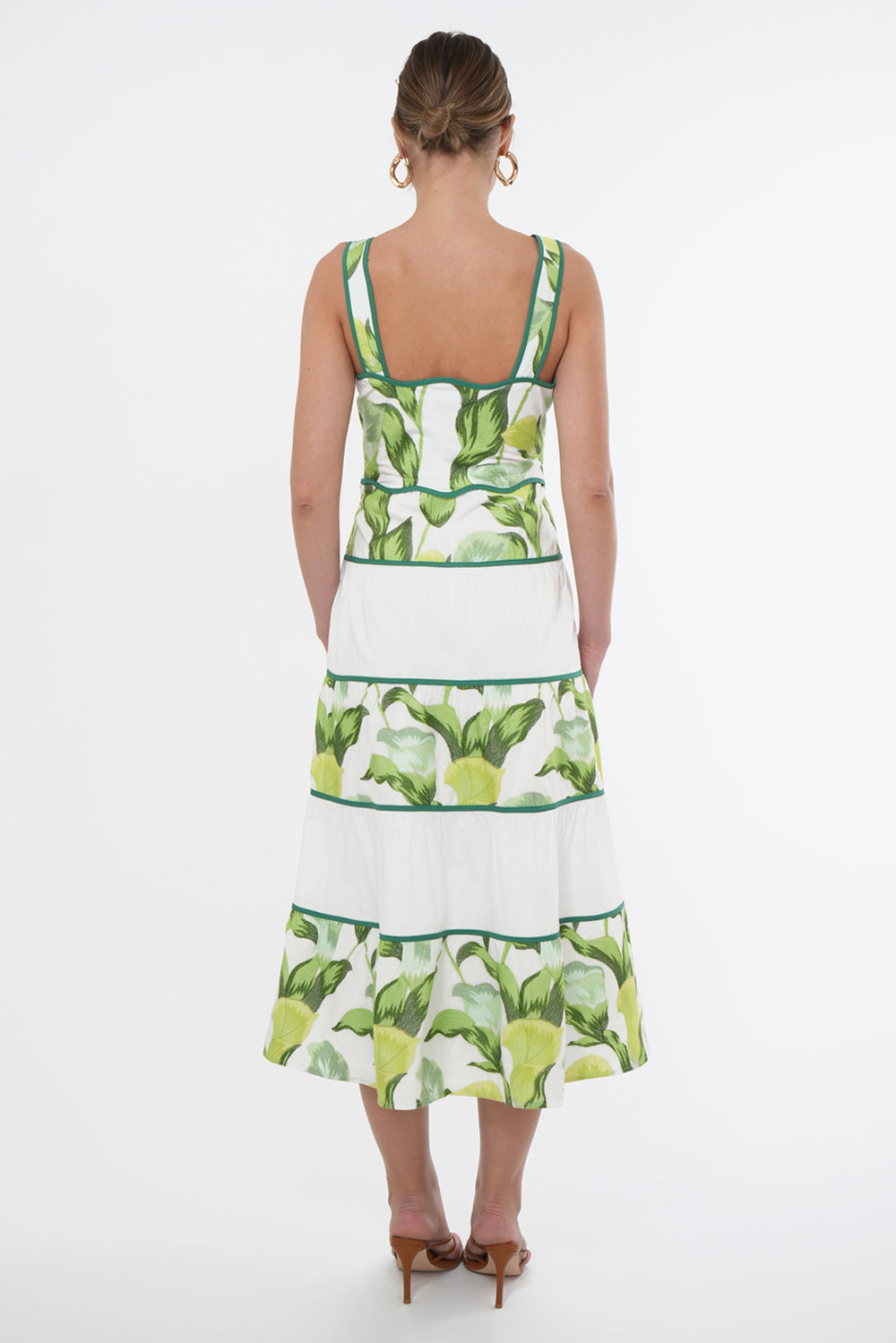 Back view of model wearing a sleeveless midi dress the dress features a green floral print on top of a white base. The neckline and waistline is scalloped. The skirt has contrasting, white tiers.